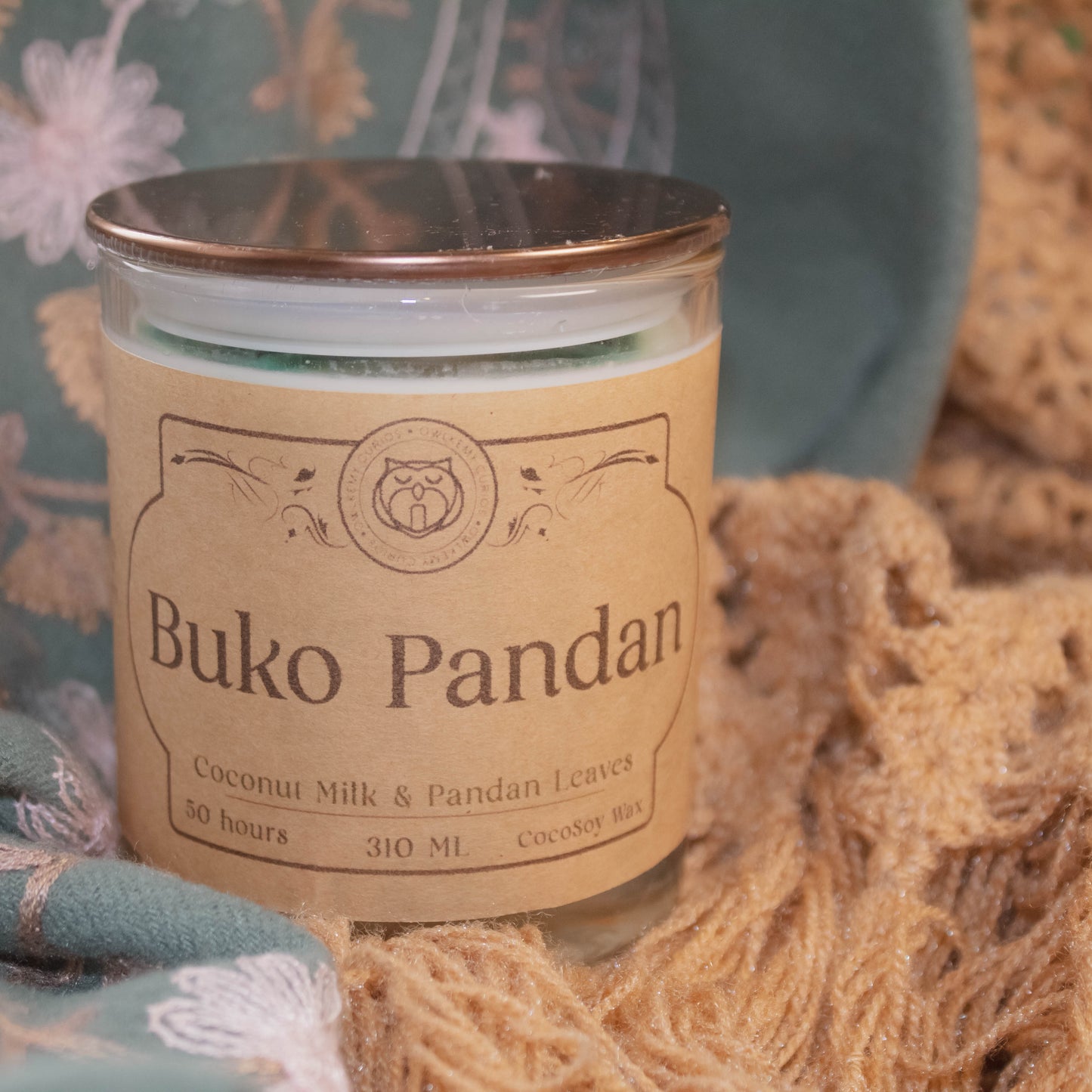 Candle jar labeled 'Buko Pandan' on a textured surface with floral and woven elements.