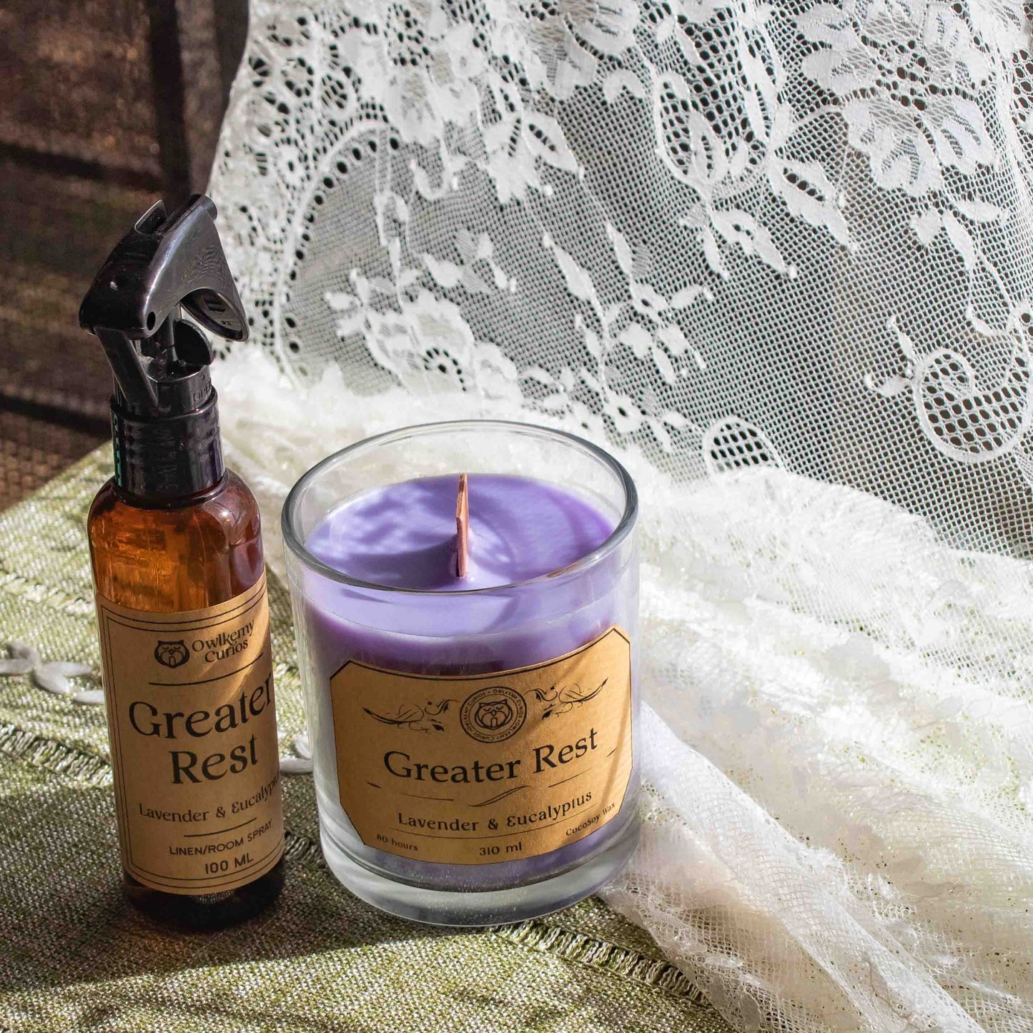 Candle and spray bottle labeled 'Greater Rest' on a textured surface with lace in the background