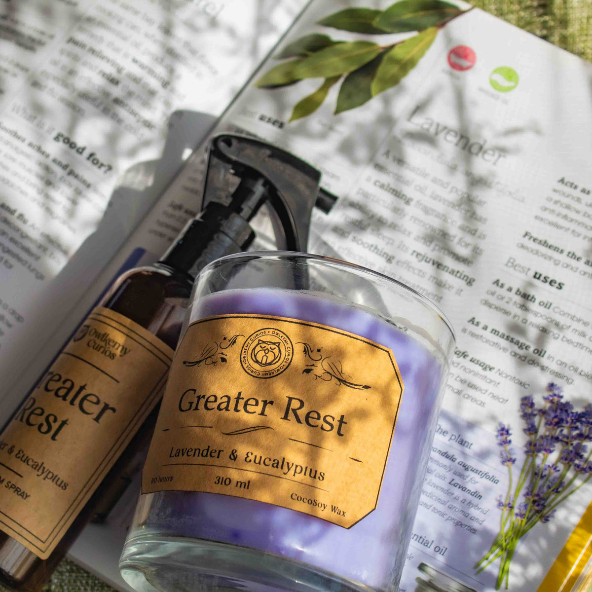 A spray bottle and a soft purple candle of 'Greater Rest' products on an open book with lavender and eucalyptus imagery.