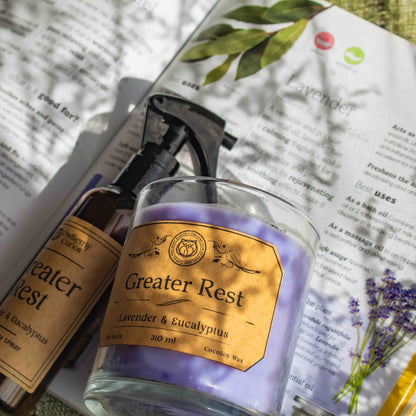 A spray bottle and a soft purple candle of 'Greater Rest' products on an open book with lavender and eucalyptus imagery.