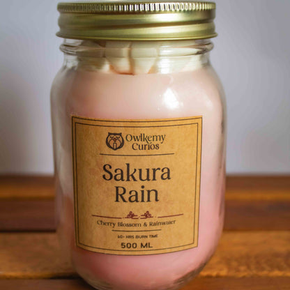 Pink candle in a glass jar with a gold lid on a wooden surface, branded 'Owlery Curios'.