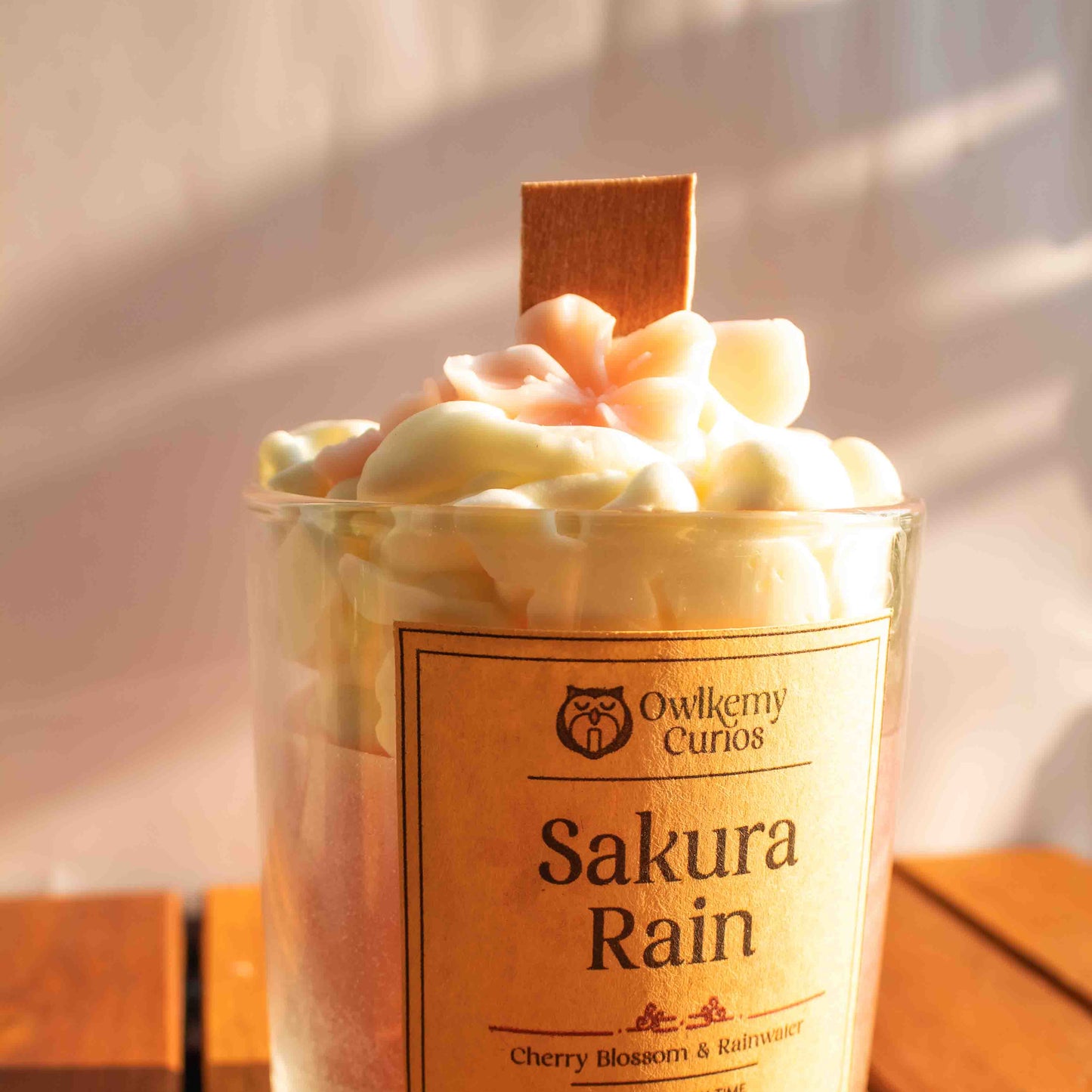 Scented candle labeled 'Sakura Rain' by Owlchemy Curios on a wooden surface.
