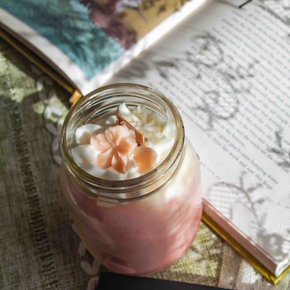 Pink candle with whipped cream-like white wax and a pink flower in a glass jar on a textured surface with books.