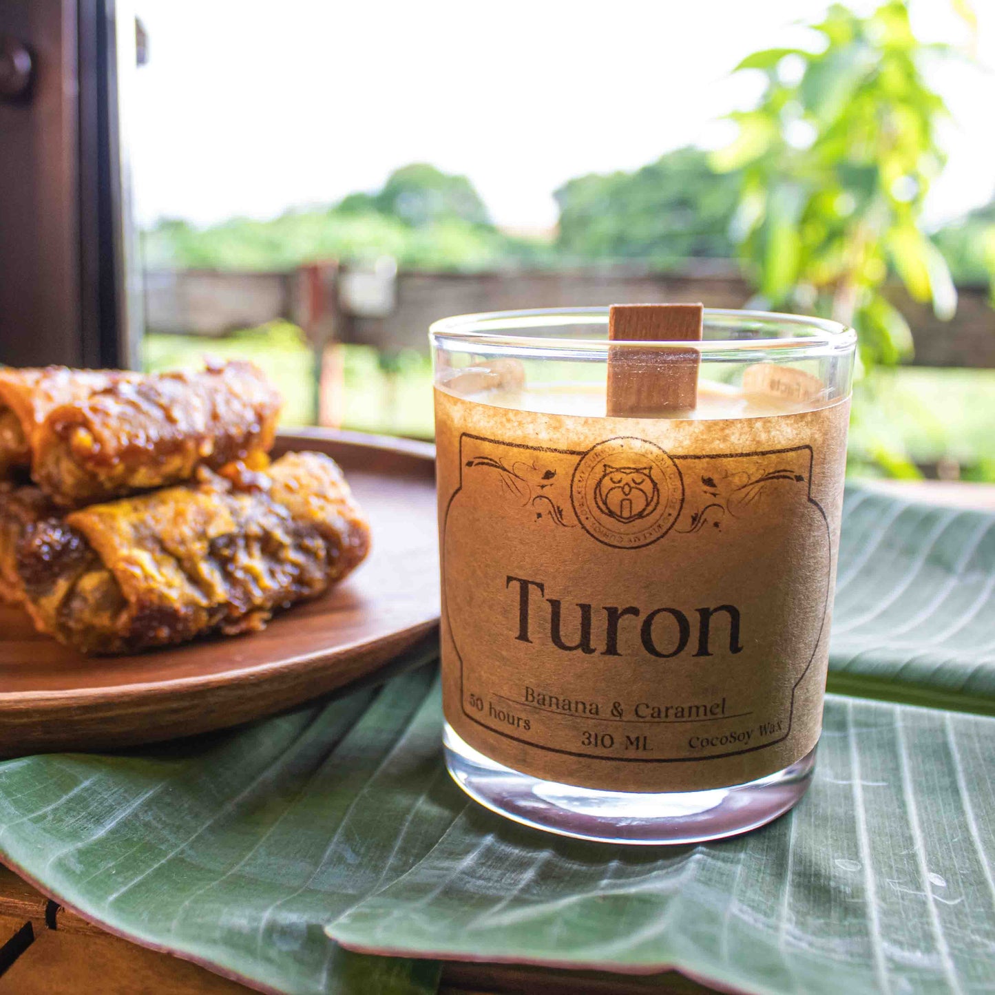Turon wrapped in banana leaves with a candle labeled 'Turon' on a leafy surface outdoors.